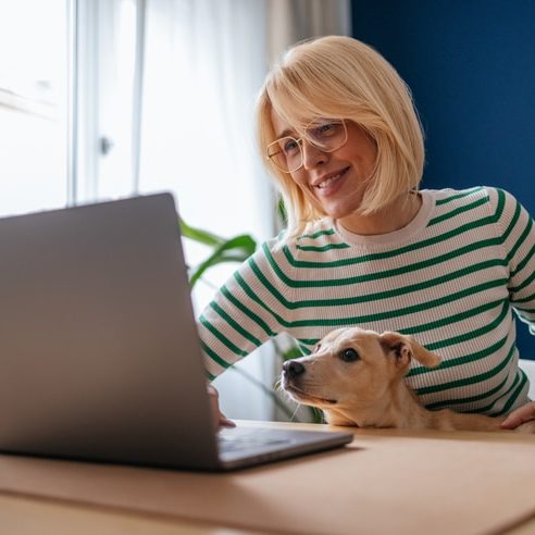 Beautiful smiling woman sitting at the desk and using her laptop computer for work or an online call. Cute yellow puppy is sitting in her lap and looking at the screen with her.